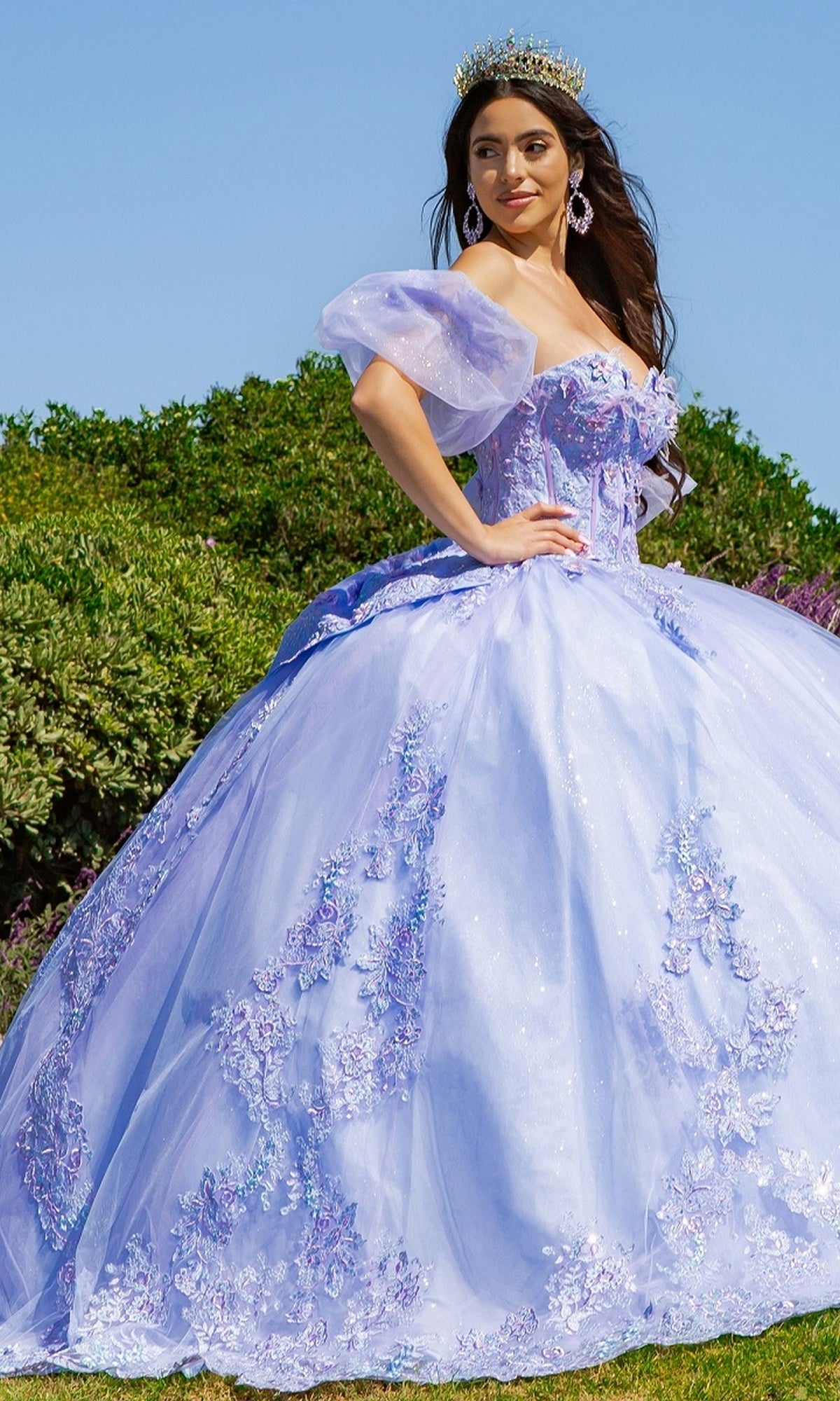 A woman stands outdoors wearing the Quinceanera Ball Gown 8115J in light purple, featuring floral embellishments and removable puff sleeves, complemented by a crown and earrings.